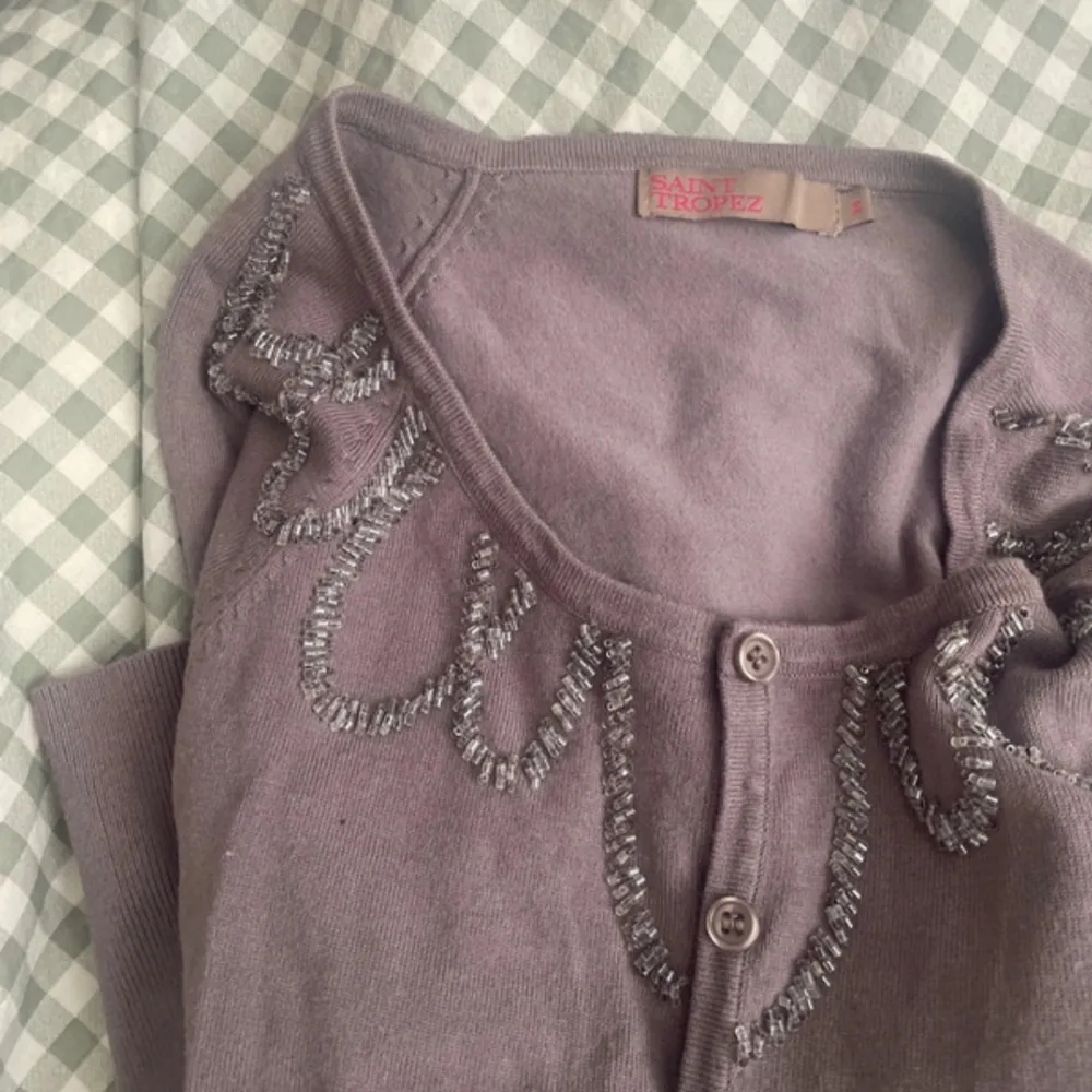 Säljer en elegant lila kofta från Saint Tropez med dekorativa pärlor längs halsringningen. Koftan har långa ärmar och knäppning framtill. Perfekt för att lägga till en touch av glamour till din outfit.. Neuletakit & Villapaidat.