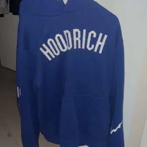 Blå hoodie från Hoodrich - Säljer en blå hoodie från Hoodrich med vit text på bröstet och ärmen. Den har en klassisk design med en stor ficka framtill och en bekväm huva. Perfekt för en avslappnad stil. Använt fåtal gånger