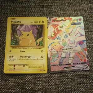 Pokémonkort: Pikachu och Sylveon - Två Pokémonkort till salu: ett klassiskt Pikachu-kort med 60 HP och attackerna Gnaw och Thunder Jolt, samt ett glittrande Sylveon VMAX-kort med 310 HP och attackerna Precious Touch och Max Harmony. Perfekt för samlare eller Pokémon-fans!