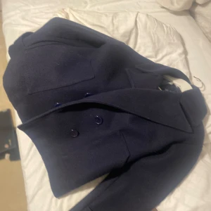 Mörkblå kappa - Snygg mörkblå kappa med dubbelknäppning och klassisk krage. Perfekt för kyligare dagar och ger en stilren look. Passar bra till både jeans och mer formella outfits. Lite nopprig