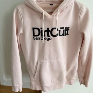 Ljusrosa hoodie från DirtCult - Säljer en ljusrosa hoodie från DirtCult med tryck 'San Diego' på framsidan. Tröjan har en klassisk känguruficka och justerbar huva med dragsko. Perfekt för en avslappnad stil.