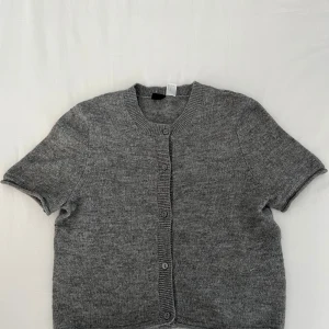 Grå kofta från H&M Divided - Säljer en stilren grå kofta från H&M Divided. Den har korta ärmar och knappar framtill. Koftan är gjord i ett mjukt material som ger en bekväm passform. Aldrig använd!! 