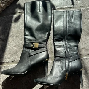 Svarta stövlar med klack och dragkedja - Snygga svarta stövlar i skinn med spetsig tå och klack. De har en dekorativ guldfärgad spänne och dragkedja på sidan för enkel på- och avtagning. Perfekta för en elegant look.