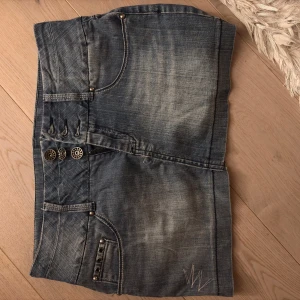 Jeanskjol med knappar - Snygg jeanskjol i blått med dekorativa knappar framtill och nitar på fickorna. Kjolen har en kort längd och är perfekt för en avslappnad stil. Den har också två bakfickor med nitar och en cool tvättad look.