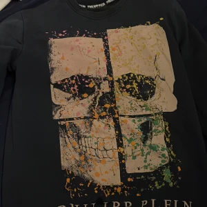 Svart tröja med dödskalletryck från Philipp Plein - Säljer en svart tröja från Philipp Plein med ett stort, färgglatt dödskalletryck på framsidan. Tröjan har långa ärmar och en rund halsringning. Perfekt för dig som gillar en edgy stil!