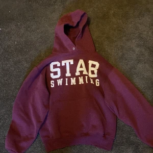 Vinröd hoodie från Jerzees - Säljer en vinröd hoodie från Jerzees med texten 'STAB SWIMMING' i vitt på framsidan. Tröjan har en klassisk känguruficka och en bekväm huva. Perfekt för kyliga dagar eller som en avslappnad vardagströja.