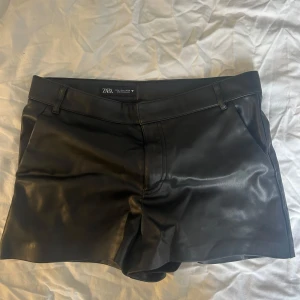 Svarta skinnshorts från Zara - Snygga svarta skinnshorts från Zara med en stilren design. De har en hög midja och är perfekta för en trendig look. Shortsen har hällor för bälte och en diskret dragkedja framtill. Supersnygga med strumpbyxor till!!!passar s-m