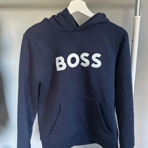 Mörkblå hoodie från BOSS - Säljer en stilren mörkblå hoodie från BOSS med vit logga på bröstet. Tröjan har en klassisk känguruficka och en bekväm huva. Perfekt för en avslappnad look.