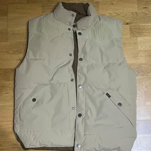 Beige och brun dunväst från Lee (Lee reversible western puffer - Väst - sand/beige - storlek M) - Säljer en snygg beige och brun dunväst från Lee i storlek M. Orginalpriset ligger på 1400 kr och är helt och hållet oanvänd. Säljer den då storleken ej passar. Västen har en quiltad design med knappar framtill. Den har praktiska fickor med knappar och en stilren krage. Perfekt för våren.  Finns absolut inga defekter med västen heller.