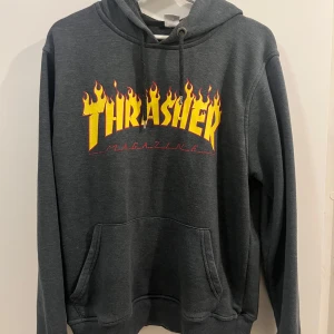 Grå hoodie från Thrasher - Säljer en grå hoodie från Thrasher med den ikoniska loggan i gult och rött på bröstet. Tröjan har en klassisk passform med känguruficka och justerbar huva. 