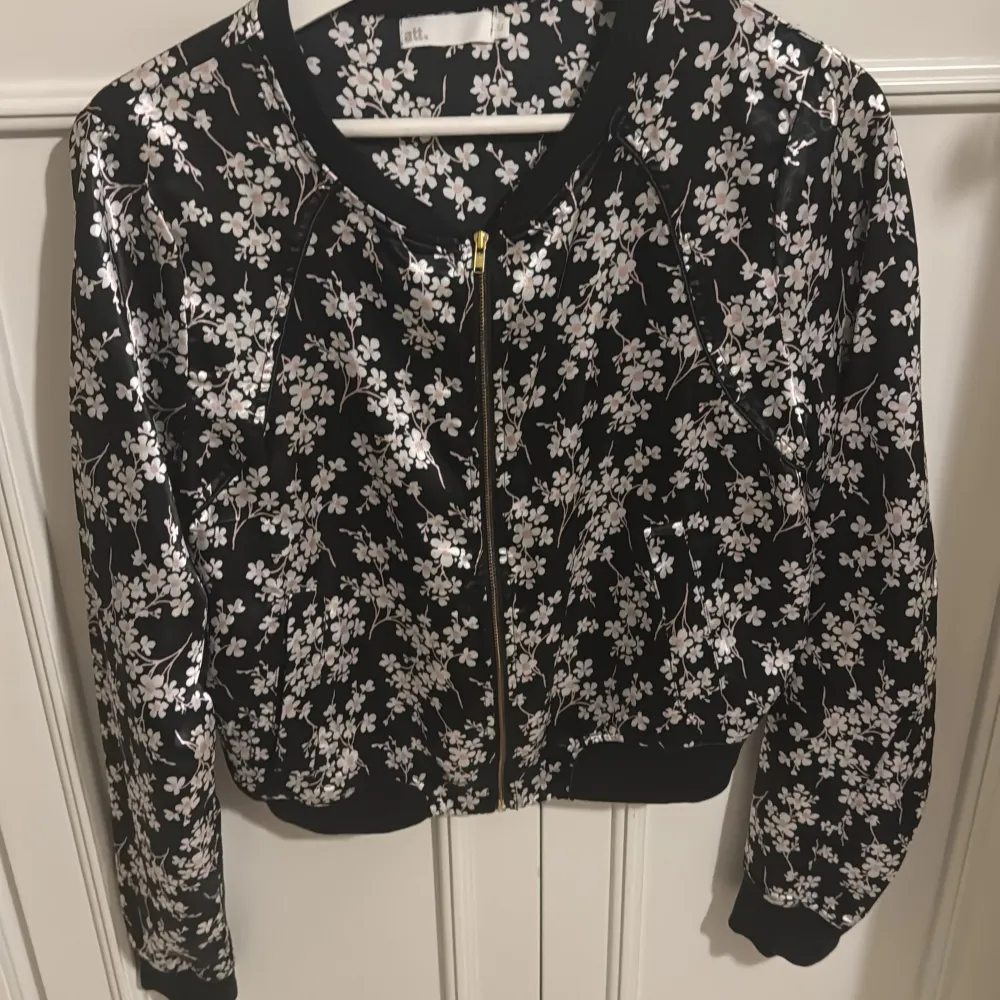Snygg svart blus från att. med ett vackert blommönster i vitt och rosa. Blusen har en dragkedja framtill och långa ärmar. Perfekt för att ge din outfit en trendig touch!. Puserot.