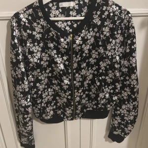 Svart blommig blus från att. - Snygg svart blus från att. med ett vackert blommönster i vitt och rosa. Blusen har en dragkedja framtill och långa ärmar. Perfekt för att ge din outfit en trendig touch!