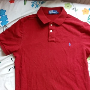 Röd pikétröja från Ralph Lauren - Säljer en klassisk röd pikétröja från Ralph Lauren med den ikoniska blå broderade loggan på bröstet. Tröjan har en krage och knappar framtill. Perfekt för en stilren look!