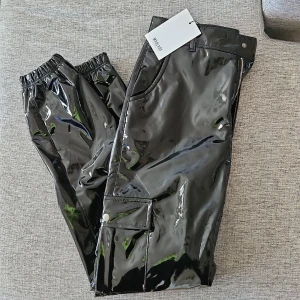 Svarta vinyl byxor från MadLady  - Snygga svarta vinyl byxor från MadLady med elastisk midja och dragkedja. Perfekta för en trendig look. Byxorna har en cool, modern stil och är lätta att matcha med olika toppar.