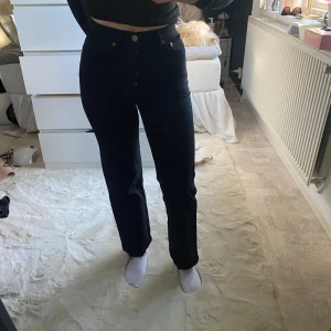 Svarta jeans med hög midja - Snygga svarta jeans med hög midja och klassisk design. De har en rak passform och är perfekta för en stilren look. Jeansen har knappar och en liten läderdetalj vid midjan.