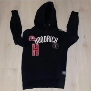 Svart hoodie från Hoodrich - Säljer en svart hoodie från Hoodrich med stora vita och röda bokstäver på framsidan. Hoodien har en justerbar huva med snören och flera märken som dekoration. Perfekt för en avslappnad stil.