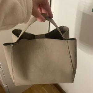 Beige axelväska med dragkedja - Snygg beige axelväska i syntetmaterial med praktisk dragkedja. Perfekt för att bära dina dagliga nödvändigheter med stil. Enkel och elegant design som passar till många olika outfits.