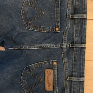 Bootcut jeans från Wrangler - Säljer ett par klassiska blå jeans från Wrangler. De är bootcut med fem fickor och bälteshällor. Jeansens bakfickor har det ikoniska V-mönstret och en läderpatch med Wrangler-loggan. Perfekta för en avslappnad stil. Säljer då de tyvärr är för korta för mig. Mått: 38 cm & 77 cm