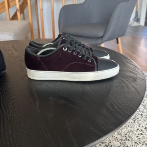 Vinröda sneakers från Lanvin - Snygga vinröda sneakers från Lanvin med svart snörning och tåhätta. Skorna har en vit sula och är tillverkade i sammet för en lyxig känsla. Perfekta för att ge din outfit en stilfull touch.