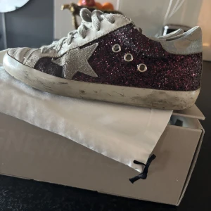 Glittriga sneakers från Golden Goose - Säljer ett par snygga sneakers från Golden Goose med glittrig vinröd yta och vita detaljer. Storlek 36. Köpta på NK i Stockholm. Ingår original dustbag och kartong. Skorna har en stjärna på sidan och snörning framtill. Säljes p g a att de blivit för små.