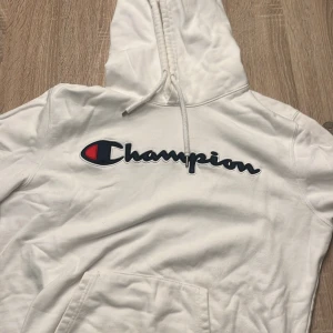 Vit hoodie från Champion - Säljer en vit hoodie från Champion med deras ikoniska logga i blått och rött på bröstet. Den har en klassisk känguruficka och justerbar huva med snören. Perfekt för en avslappnad stil.