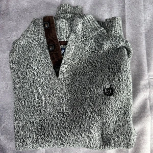 Grå stickad tröja från Chaps - Säljer en snygg grå stickad tröja från Chaps i storlek M. Tröjan har en stilren design med bruna detaljer och knappar vid halsen. Perfekt för kyligare dagar med sin mjuka bomullskvalitet.