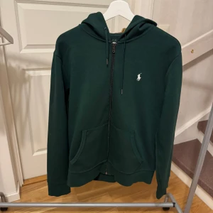 Mörkgrön hoodie från Ralph Lauren - Säljer en snygg mörkgrön hoodie från Ralph Lauren med dragkedja och klassisk logga på bröstet. Tröjan har långa ärmar och en praktisk huva med snörning. Perfekt för en avslappnad stil. Storlek: M Nypris: 1700kr