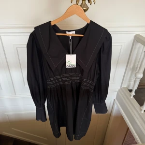 Svart klänning från Ganni - Säljer en elegant svart klänning från Ganni med kontrastsömmar och en unik krage. Klänningen har långa ärmar och en stilren design som passar perfekt för olika tillfällen. Perfekt för dig som vill ha något stilrent och modernt i garderoben.