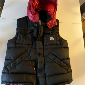 Moncler väst - Snygg dunväst från Moncler i svart och rött, storlek 3. En knapp saknas och därmed billigt pris. Har även kvitto. Köpt från resellsida. Tveka inte om ni har frågor.