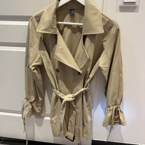 Beige trenchcoat från Shein - Snygg beige trenchcoat från Shein i storlek XS. Jackan har ett klassiskt snitt med knappar och ett bälte i midjan. Ärmarna har knytband för extra stil. Perfekt för vår och sommarkvällar!