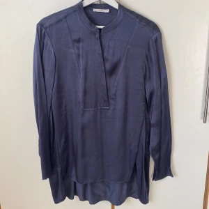 Åskblå lång blus från Wera - Snygg åskblå blus från Wera i ett mjukt och blankt material med ett tungt skönt fall. Spännande design med en diskret omlottknäppning framtill och en avslappnad passform. Längd fram 70 cm, längd bak 82 cm. Sprund i sidorna. 