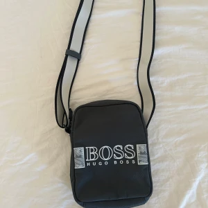 Svart axelväska från Hugo Boss - Snygg svart axelväska från Hugo Boss med justerbar axelrem. Väskan har en stilren design med vit logotyp på framsidan och är perfekt för att bära med sig det nödvändigaste. Praktisk och enkel att matcha med olika outfits. Knappt använd. Nypris ca 1100kr. Pris kan diskuteras