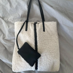 Vit shoppingväska från Carin Wester - Säljer en stilren vit shoppingväska från Carin Wester med svarta detaljer. Väskan har en mjuk, pälsliknande yta och svarta handtag. Perfekt för att bära med sig allt man behöver under dagen.