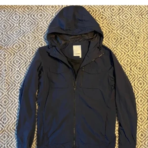 Mörkblå j lindeberg vindjacka med huva - Säljer en stilren mörkblå vindjacka med huva. Jackan har dragkedja framtill och två bröstfickor med dragkedjor. Perfekt för blåsiga dagar och har en enkel design som passar till det mesta.