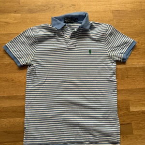 Pikétröja från Polo Ralph Lauren - Säljer en stilren randig pikétröja från Polo Ralph Lauren i blått och vitt. Tröjan har en klassisk krage och knappar framtill. Den är tillverkad i 100% bomull och har en broderad grön logotyp på bröstet. Storlek: S Skick: 8/10
