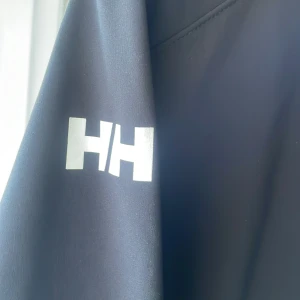Svart vindjacka från Helly Hansen - Säljer en stilren svart vindjacka från Helly Hansen. Jackan har en dragkedja framtill och en praktisk huva. Perfekt för blåsiga dagar och har en logga på ärmen. Passar bra för utomhusaktiviteter.