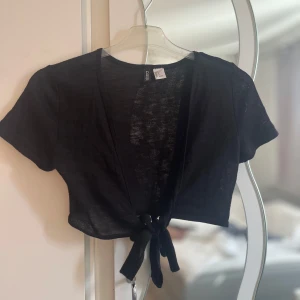 Svart bolerotopp från H&M - Säljer en svart bolerotopp från H&M. Toppen är kortärmad och har en knytning framtill. Perfekt för att lägga till en extra touch till din outfit. Endast använd 1 gång.💕