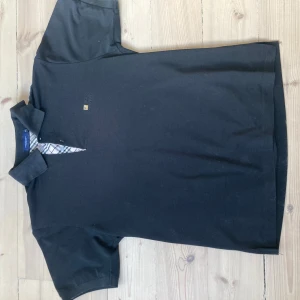 Svart pikétröja från Burberry - Snygg svart pikétröja från Burberry med klassisk krage och knappslå. Tröjan har ett diskret broderat Burberry-märke på bröstet och en stilren rutig detalj på insidan av kragen. Perfekt för en stilren look.