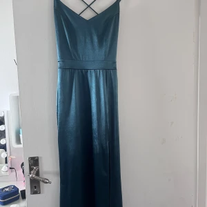 Mörkgrön långklänning med spetsdetaljer -  Mörkgrön balklänning i satin med smala axelband och v-ringning. Klänningen har spetsdetaljer i ryggen och en slits fram. Passar jättebra till bal, endast använd 2 gånger. 