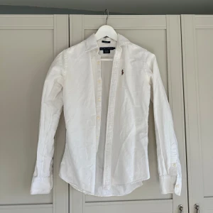 Vit skjorta från Ralph Lauren - Säljer en klassisk vit skjorta från Ralph Lauren i slim fit. Skjortan har långa ärmar och knappar framtill. Den är dekorerad med Ralph Laurens ikoniska broderade logotyp på bröstet. Perfekt för en stilren look.