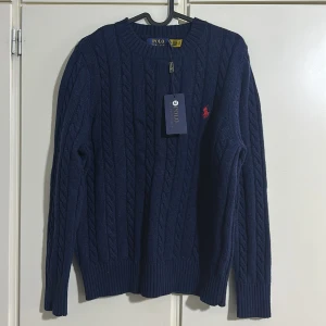 Mörkblå stickad tröja från Ralph Lauren - Säljer en stilren mörkblå stickad tröja från Ralph Lauren med det klassiska röda logotypbroderiet på bröstet. Tröjan har en kabelstickad design och långa ärmar, perfekt för kyligare dagar.