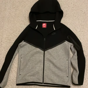 Svart och grå hoodie från Nike - Säljer en snygg svart och grå hoodie från Nike med dragkedja och luva. Jackan har en stilren design med kontrasterande färger och är perfekt för en sportig look. Den har långa ärmar och en liten Nike-logga på bröstet. Perfekt för kyliga dagar!