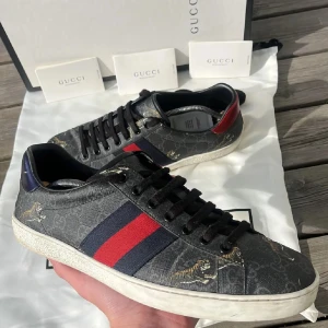 Gucci sneakers med tigertryck - Snygga Gucci sneakers med ett unikt tigertryck på en grå bas. Skorna har de ikoniska röd-blå ränderna på sidan och en kontrasterande röd häl. Perfekta för att ge din outfit en lyxig touch.