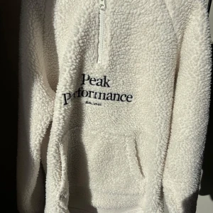 Vit fleece hoodie från Peak Performance - Mysig vit fleece hoodie från Peak Performance med halv dragkedja och broderad logga på framsidan. Perfekt för kyliga dagar med sin mjuka och varma känsla. Den har en praktisk ficka framtill och långa ärmar. 