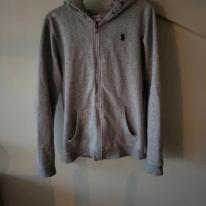 Grå hoodie från U.S. Polo Assn. - Säljer en stilren grå hoodie från U.S. Polo Assn. med dragkedja och klassisk logga på bröstet. Perfekt för en avslappnad look. Den har en bekväm passform och praktiska fickor framtill. Det finns ett märke som visas på bild 4..storleken är barn
