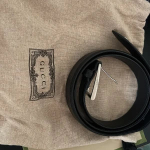 Svart bälte från Gucci - Säljer ett stilrent svart bälte från Gucci med en elegant silverfärgad spänne. Bältet kommer i sin originalförpackning med en snygg grön låda och en tygpåse med Gucci-logga. Perfekt för att ge en lyxig touch till din outfit. (Orginal pris 3000+) om du har några frågor skicka DM! Ps 115cm i längden 