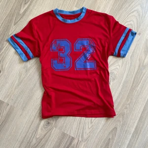 Röd t-shirt med blå detaljer och nummer 32 - Säljer en cool röd t-shirt med blå detaljer och ett stort nummer 32 tryckt på framsidan. T-shirten har korta ärmar med blå ränder.