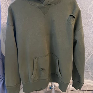Grön hoodie med slitna detaljer - Säljer en grön hoodie med slitna detaljer för en cool och avslappnad look. Tröjan har en klassisk känguruficka och ribbade muddar vid ärmslut och nederkant. Perfekt för en casual stil. Fläck på magen.