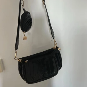 Svart axelväska med krokodilmönster - Snygg svart axelväska med krokodilmönster och gulddetaljer. Väskan har en justerbar axelrem och en praktisk dragkedja. Perfekt för att bära dina nödvändigheter med stil.