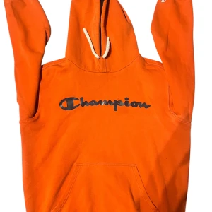 Orange hoodie från Champion - Säljer en cool orange hoodie från Champion med klassisk logga på bröstet. Den har en känguruficka och justerbar huva med vita snören. Perfekt för en avslappnad stil.